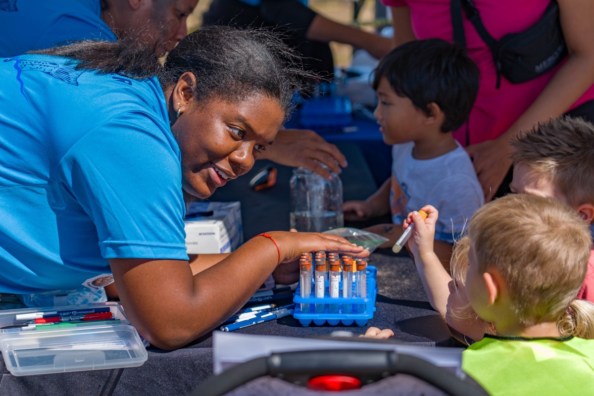 A smiling woman in a blue shirt shows young children a rack of water sample tubes at a hands-on water quality table at the Waterways Festival.